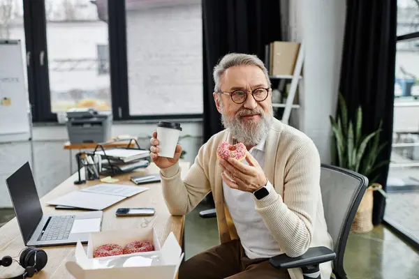 Senior man enjoys a donut and coffee at his desk, surrounded by work materials. — Stock Photo