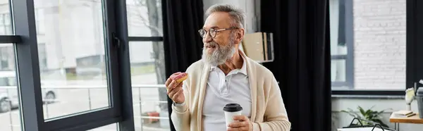 Ein reifer Mann mit grauen Haaren hält eine Kaffeetasse und ein Gebäck in der Hand und lächelt in einem stilvollen Büro. — Stockfoto