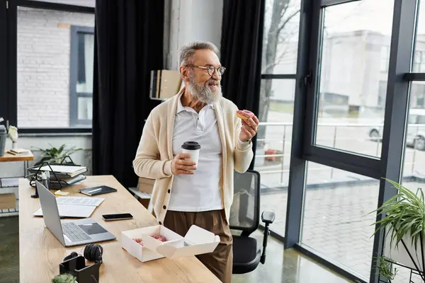 Handsome senior man stands in a contemporary office, sipping coffee and holding a pastry. — Stock Photo