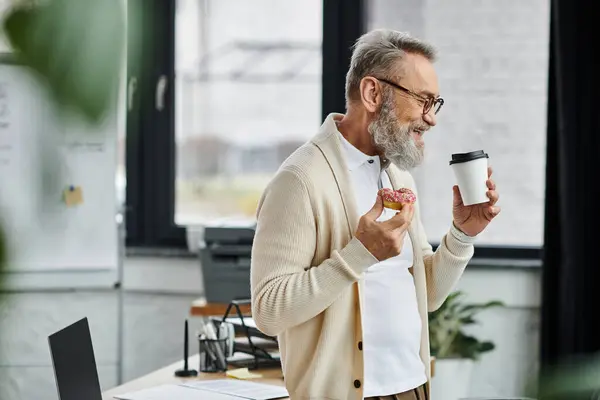 Senior man smiles while holding a coffee cup and a donut in a modern workspace. — Stock Photo