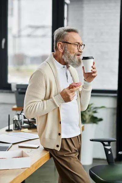 Handsome senior man leans against a desk savoring a donut and sipping coffee in a bright office. — Stock Photo