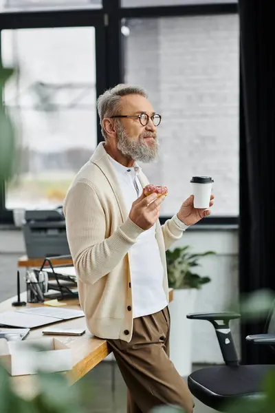 Un bell'uomo maturo assapora una pasticceria e un caffè mentre si trova casualmente in un ufficio. — Foto stock