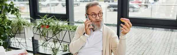 A mature handsome man with a beard is speaking on the phone and holding a donut. — Stock Photo