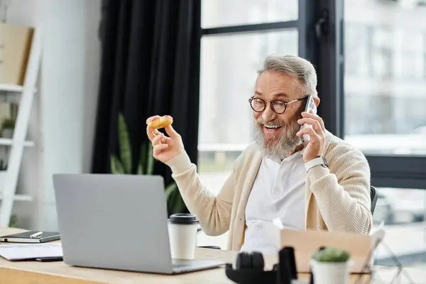 Handsome senior man smiles happily on a phone call while holding a donut at his desk. — Stock Photo
