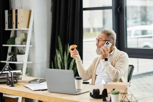 Handsome older man multitasks at his desk, enjoying a donut and talking on the phone. — Stock Photo