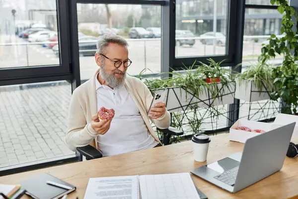 Un uomo barbuto alla scrivania si gode una ciambella mentre usa il suo smartphone in un ufficio moderno. — Foto stock