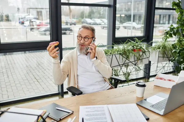 Uomo impegnato in una conversazione telefonica mentre tiene una ciambella colorata e seduto a una scrivania. — Foto stock