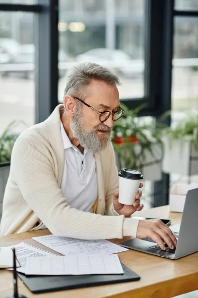 Senior man with a beard engaged in work on a laptop while holding a coffee cup. — Stock Photo