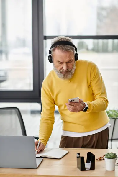 Handsome senior man wearing headphones engages with his smartphone at a desk. — Stock Photo