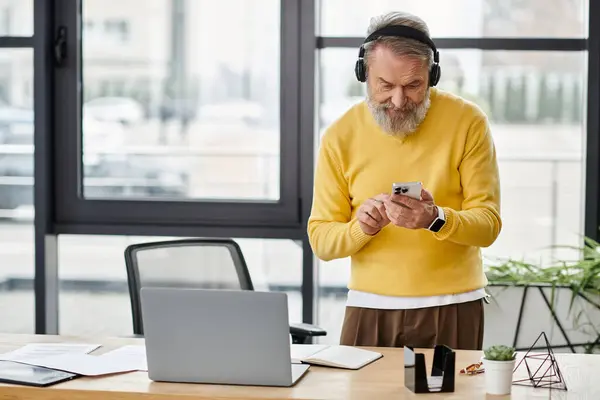 Handsome senior man with headphones checks his smartphone while standing at a desk. — Stock Photo