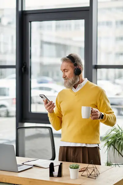 Un hombre guapo maduro con barba escucha música en los auriculares mientras sostiene una taza de café. - foto de stock