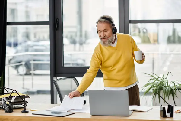 Uomo in un maglione giallo esamina i documenti alla sua scrivania mentre tiene in mano una tazza di caffè. — Foto stock
