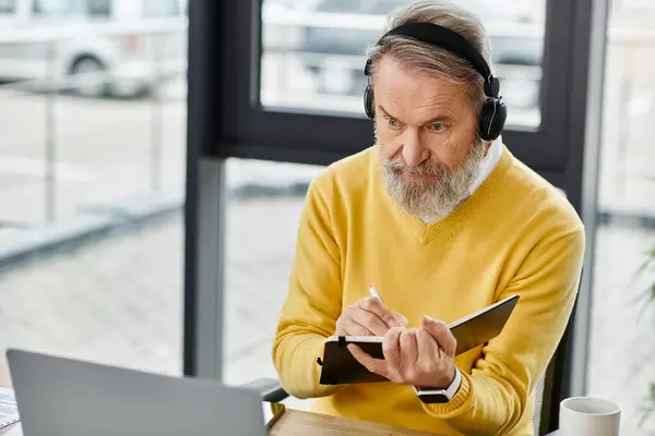 Mature man wearing headphones writing notes while focused on a laptop in an office. — Stock Photo
