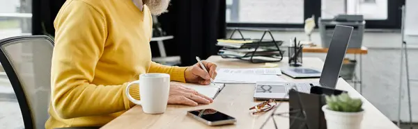 Handsome senior man writes notes while working at his desk with a cup of coffee nearby — Foto stock