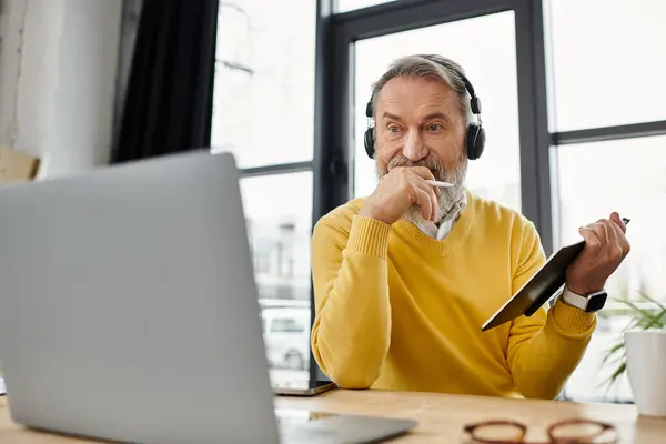 Bello uomo anziano si concentra sul suo computer portatile, prendendo appunti durante l'ascolto attraverso le cuffie. — Foto stock