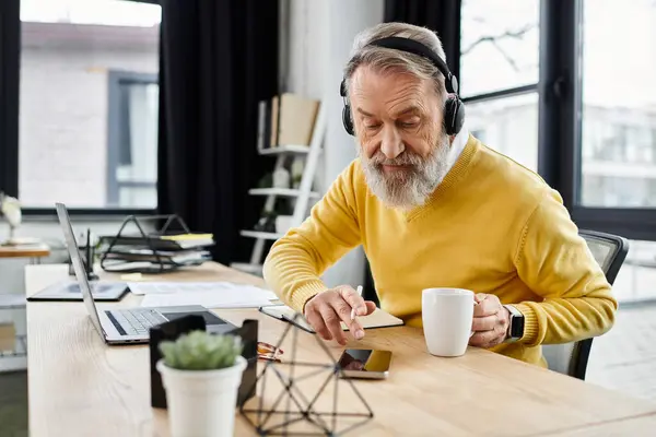 Handsome senior man in yellow sweater listens to music while working at his desk. — Stock Photo
