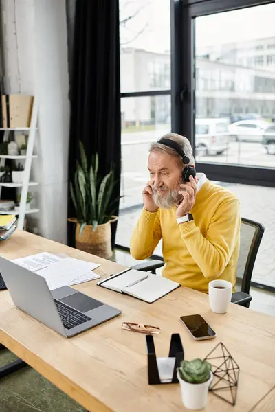 A mature handsome man wearing a yellow sweater engages in work while seated at a desk with a laptop. — Stock Photo
