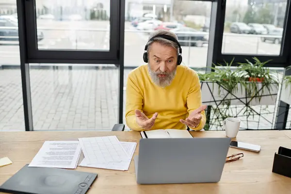 Handsome senior man wearing headphones gestures while discussing in virtual meeting at office. — Stock Photo