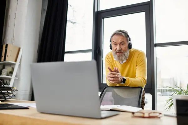 Hombre mayor guapo con auriculares utiliza activamente el ordenador portátil en un espacio de trabajo brillante. - foto de stock