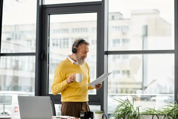 Handsome senior man standing in bright office, sipping coffee and examining paperwork thoughtfully. — Stock Photo