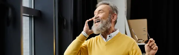 A mature man smiles broadly while talking on his phone, holding glasses with one hand. — Stock Photo
