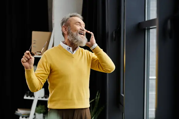 Bell'uomo anziano vestito con un maglione giallo sorride mentre parla al telefono a casa. — Foto stock