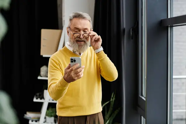 Uomo maturo in un maglione giallo utilizzando il suo smartphone mentre in piedi in una stanza elegante — Foto stock