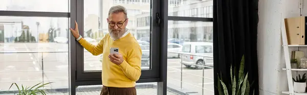Ein älterer Herr im gelben Pullover checkt sein Handy, während er nach draußen in Richtung Stadtansicht blickt.. — Stockfoto