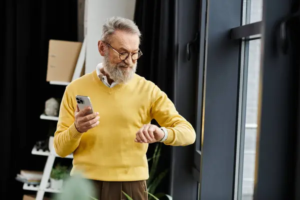 Bello uomo anziano guarda il suo smartwatch mentre tiene uno smartphone in casa. — Foto stock