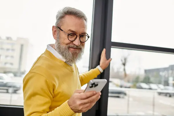 Senior man with glasses smiles while checking his smartphone by a large window. — Stock Photo