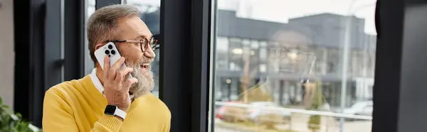 Mature man engages in a cheerful conversation on his smartphone near a bright window. — Stock Photo