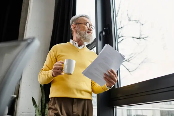 Handsome senior man wearing a sweater holds a cup and papers while looking outside. — Stock Photo