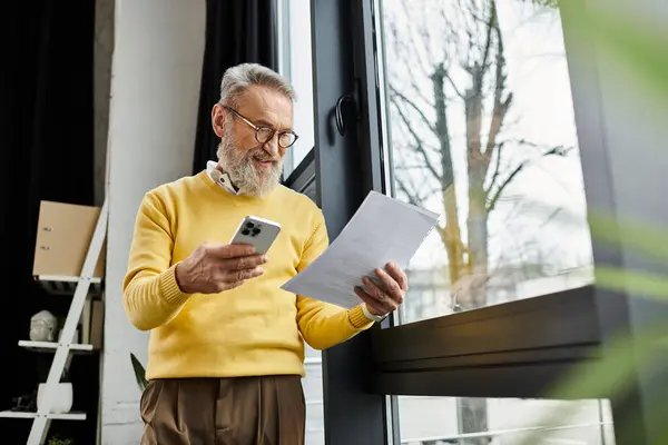 Senior man in a yellow sweater reviews documents while checking his smartphone. — Stock Photo