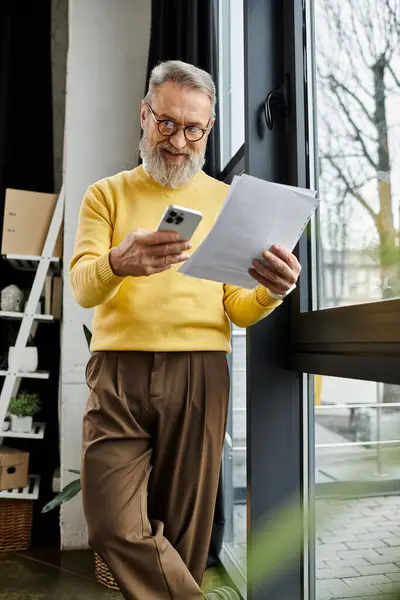 A mature handsome man is reading documents while checking his smartphone by the window. — Stock Photo