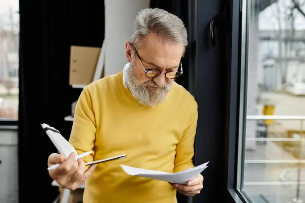 Homme mûr en pull jaune passe en revue les documents importants tout en se tenant près d'une fenêtre. — Photo de stock