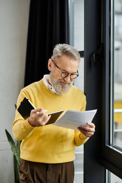 Handsome senior man in a yellow sweater engaged in reviewing papers by the window. — Stock Photo