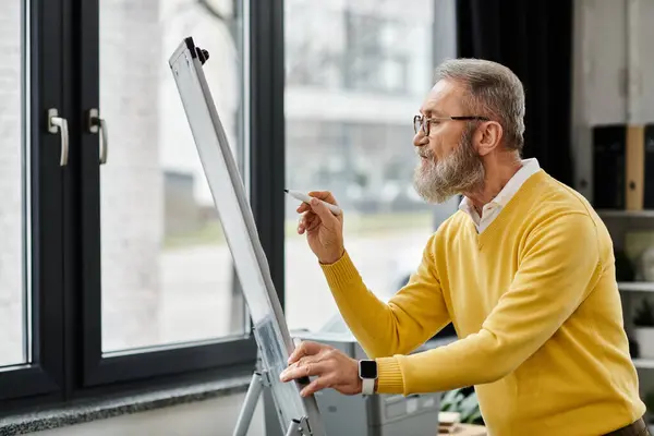 Handsome senior man focuses on drawing at an easel near a sunny window in a studio. — Stock Photo