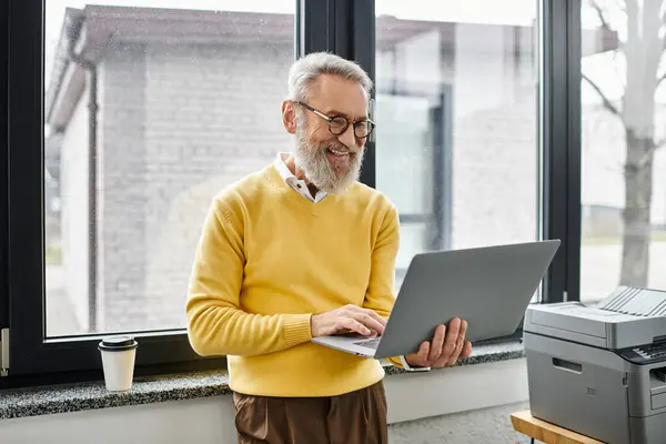 A mature handsome man smiles while using a laptop near a window in a bright office. — Stock Photo