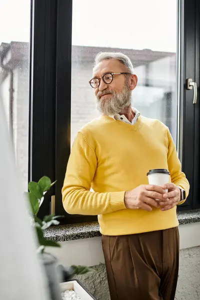 Mature man holds a coffee cup, gazing out of the window, dressed casually in a yellow sweater. — Stock Photo