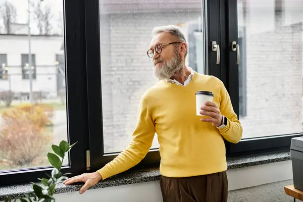 Ein reifer Mann mit Bart hält eine Kaffeetasse in der Hand, während er warm angezogen aus dem Fenster blickt.. — Stockfoto