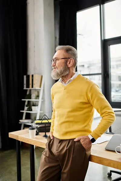 A mature handsome man leans casually against a desk in a modern office while gazing out the window. — Stock Photo