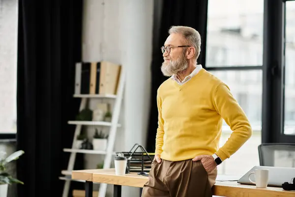 A mature handsome man wearing a yellow sweater stands by a desk in a modern office setting. — Stock Photo