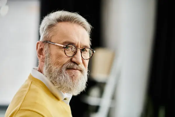 A mature handsome man with a beard and glasses smiles softly while dressed in a yellow sweater. — Stock Photo