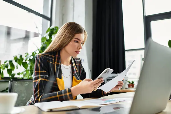A millennial businesswoman reviews documents while using her smartphone in a bright office. — Stock Photo