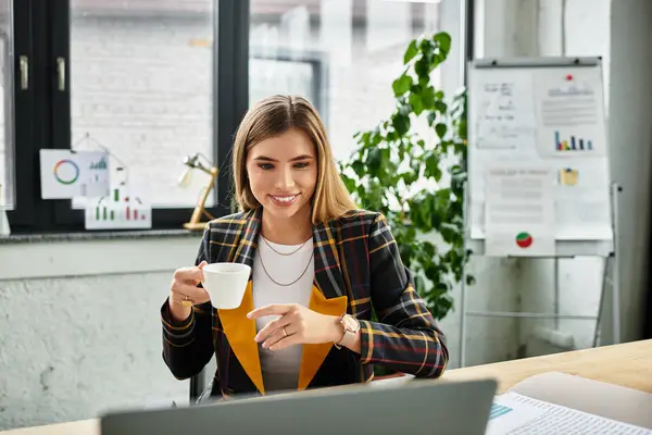 A stylish young woman with a checkered blazer smiles as she sips coffee and works at her desk. — Stock Photo