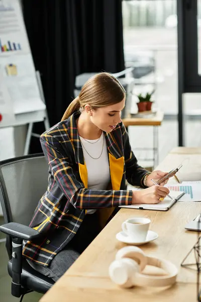 Young woman in a stylish blazer writes notes while using her smartphone in an office. — Stock Photo