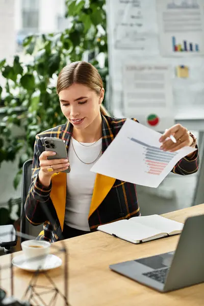 Engaging in work, a young woman reviews data on her phone while examining charts at her desk. — Stock Photo