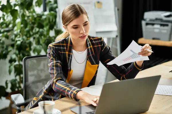 Attractive young woman focuses on her laptop while reviewing documents in a contemporary workspace. — Stock Photo
