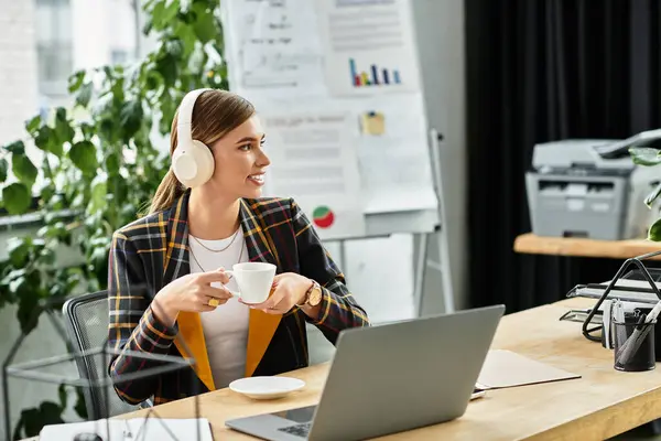 En una oficina moderna, una mujer joven bebe café, usando una chaqueta a cuadros de moda y auriculares. - foto de stock