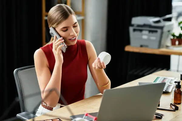 Young woman having phone call while holding face cream next to laptop — Stock Photo